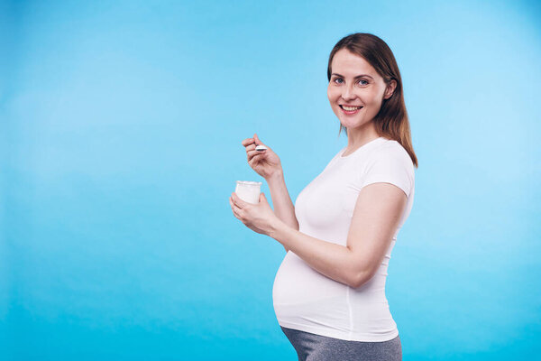 Cheerful and healthy young pregnant woman looking at you while eating greek yoghurt for breakfast in isolation on blue background