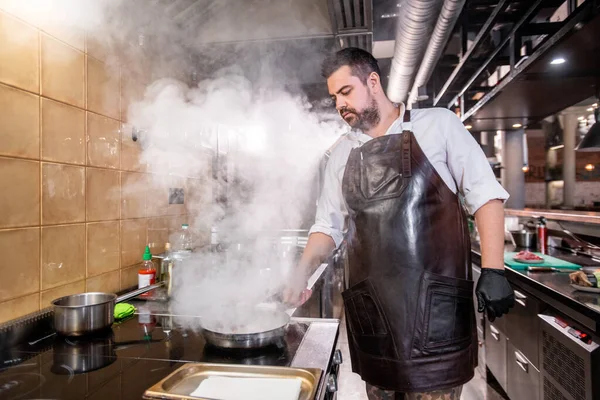 ストーブの上に立つエプロンで真剣なひげそりのシェフと鍋に食材を煮込み 日本の上の蒸気 — ストック写真