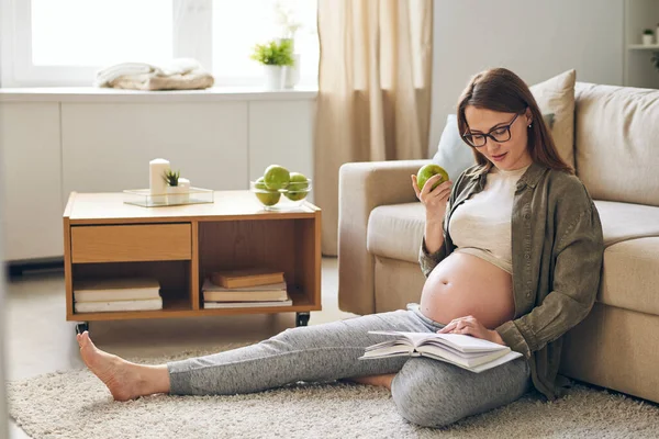 年轻的怀孕妇女穿着便服 边看书边吃着青苹果 坐在家里的沙发上 — 图库照片