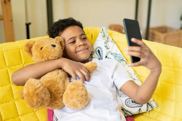 Akıllı Telefonuyla Oyuncak Ayısını Kucaklayan Sarı Koltukta Oturup Selfie Çeken — Stok fotoğraf