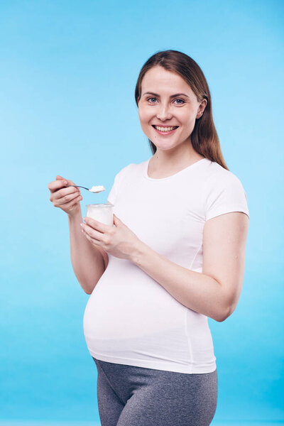 Pretty pregnant woman in activewear holding small plastic jar with bio yoghurt while eating it in front of camera in isolation