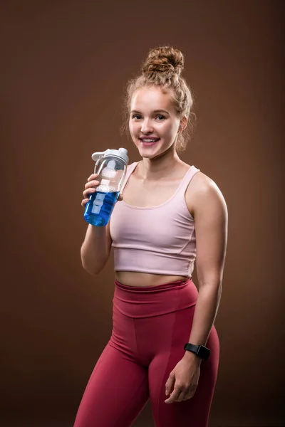 ジムでのトレーニング後に休憩しながら さわやかな飲み物とプラスチックボトルを保持歯の笑顔を持つ若い陽気なスポーツ女性 — ストック写真