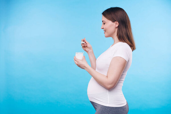 Profile view of young healthy pregnant female in white shirt eating yoghurt from small plastic cup while standing on blue background