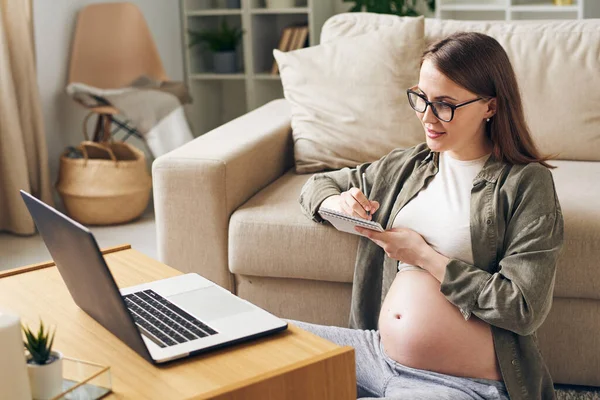 在网上医疗会诊期间 快乐的孕妇坐在笔记本电脑前的沙发上 在笔记本上做笔记 — 图库照片