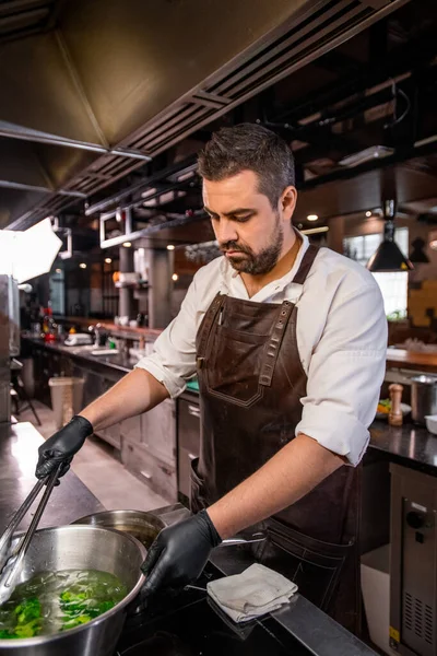 台所で鍋にストーブと沸騰ブロッコリーに立ってエプロンで忙しいひげそりのシェフ — ストック写真