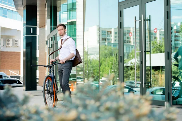 Contemporary young office manager in formalwear standing by door of business center while going to go back home by bicycle