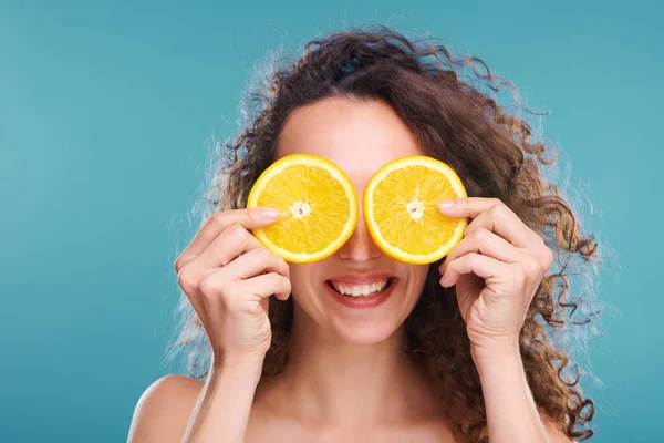 年轻快乐的卷发女子 两片新鲜多汁的橙子在她的眼睛旁 孤立地站在蓝色的背景上 — 图库照片