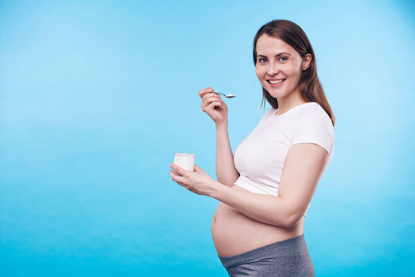 Young smiling future mother enjoying yoghurt while eating it from small plastic container over blue background with copyspace on the left