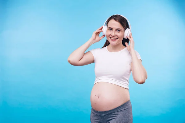 年轻快乐的怀孕女性 面带微笑地看着你 在摄像机前的耳机里欣赏她最喜欢的音乐 — 图库照片
