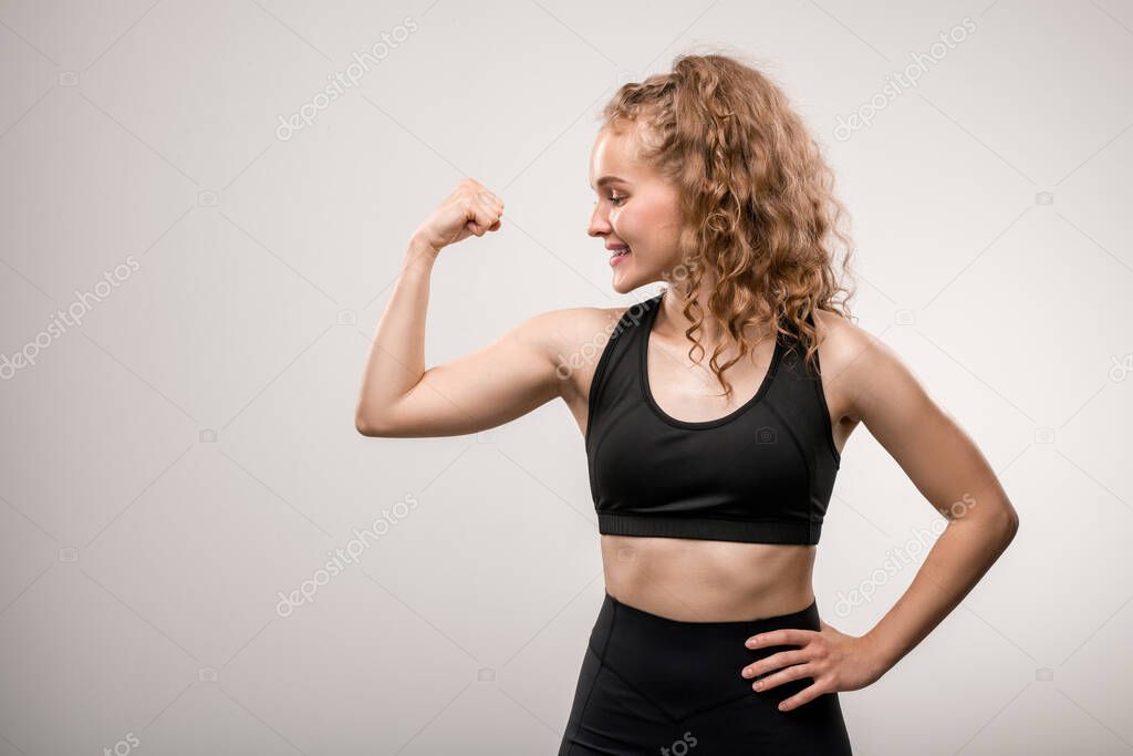 Young cheerful sportswoman with long blond curly hair keeping one hand on waist while showing her physical strength in front of camera