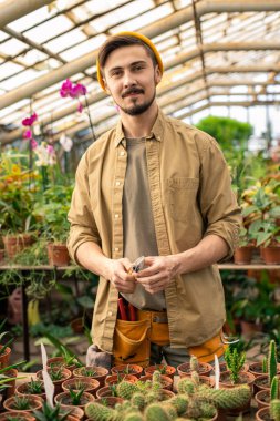 Kendine güvenen genç sakallı, günlük gömlekli, portakallı kürek taşıyan bir sera işçisinin portresi.
