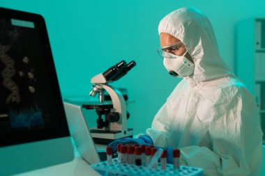 Odaklanmış koronavirüs araştırmacısı kanı analiz ediyor ve tıp laboratuarında dizüstü bilgisayar kullanarak hastalıklar hakkında notlar alıyor.