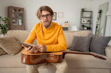 Genç başarılı gündelik kıyafet müzik öğretmeni kanepeye oturmuş online ders sırasında not defterine notlar yazarken sana bakıyor.