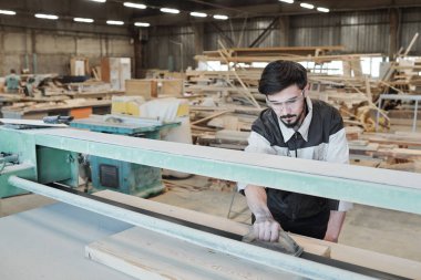 İş elbisesi içinde genç bir adam tezgahın üzerinde eğiliyor ve fabrikada çalışırken ahşap bir aletle çalışma yüzeyini düzleştiriyor.