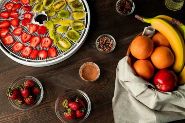 Ahşap mutfak masasındaki taze meyve çeşitliliğinin en üst görüntüsü dilimlenmiş kivi ve kurutma tepsisindeki çilekler ve aromatik baharatlar da dahil.