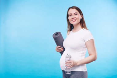 Bir şişe su ve hasırla sana gülümserken mavi arka planda duran mutlu, genç, aktif, hamile bir kadın.