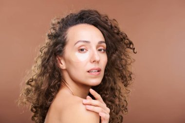 Muhteşem siyah ve kıvırcık saçlı kadın. Gözlerinin altında gençleştirici bantlar var. Kameranın önünde dururken çıplak omzuna dokunuyor.