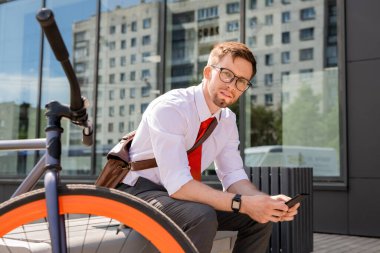 Genç işadamı iş gününden sonra bankta otururken ve şehir ortamında akıllı telefon kullanırken sana bakıyor.