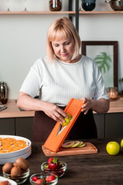 Olgun ev hanımı mutfak masasında taze yeşil armut keserken aynı zamanda ev ortamında kış için çeşitli meyveler hazırlıyor.