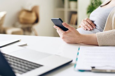 Ofis ortamında dizüstü bilgisayarın önünde otururken akıllı telefon kullanan hamile bir iş kadını.