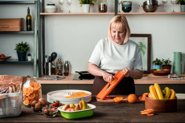Sarışın, güzel ev hanımı mutfak masasının yanında duruyor ve taze portakalları kivi, çilek, armut ve muzla kaseler arasında kesiyor.
