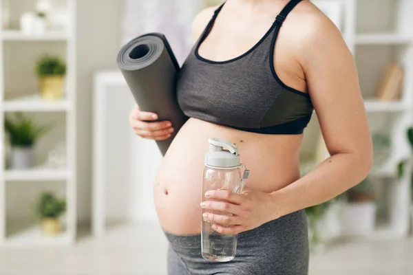 年轻而活跃的孕妇 拿着滚动的垫子和一瓶清澈的水 站在镜头前进行体育锻炼 — 图库照片