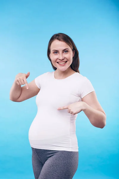 穿着运动服的快乐的孕妇指着她的腹部 表示在蓝色背景下期待孩子的快乐 — 图库照片