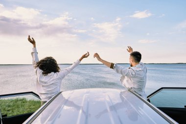 Arabalarının yanında duran, kapıları açık duran ve ellerini kaldırarak özgürlüğün sevincini ifade eden gömlekli neşeli çiftin arka görüntüsü.