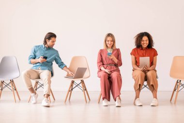 Kültürler arası iki kadın milenyum sandalyede oturup mobil cihazlar kullanırken akıllı telefonlu genç adam dizüstü bilgisayarın anahtarını itiyor.