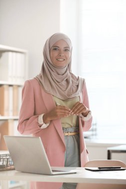 Kafe masasında çalışan, gülümseyen Müslüman bir iş kadını.