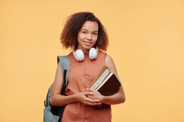 Boynunda kulaklıklarla gülümseyen Afro-Amerikalı bir kızın portresi. Sırtında parlak bir arka planda kitap ve çantasıyla.