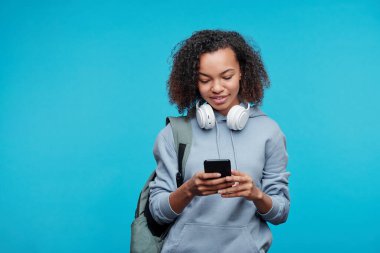 Ensesinde kablosuz kulaklıkları olan Afro-Amerikan öğrenci kız telefon mesajlarını mavi arka planla karşılaştırıyor.