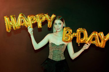 Sarışın, zarif, şık elbiseli, elinde altın harflerle balon şekilli bir kadın doğum gününde birini tebrik ediyor.