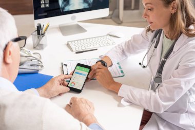 Whitecoat 'taki genç beslenme uzmanı danışma sırasında yeni geliştirilmiş sağlık göstergeleriyle kıdemli hasta akıllı telefonunu ekranında gösteriyor.