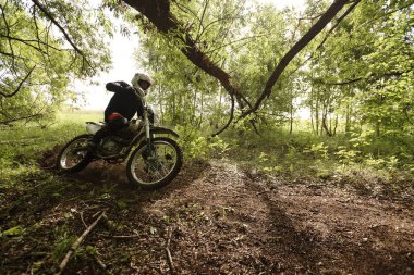 Kasklı sporcu motosiklet kullanıyor ve orman yolunu geçerken sırayla motosikletten eğiliyor.