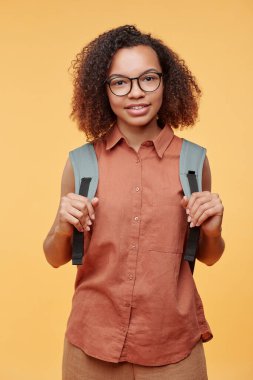 Gözlüklü gülümseyen Afro-Amerikan lise öğrencisinin portresi sarı arka planda sırt çantası kayışları taşıyor.