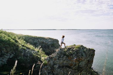 Genç bayan yürüyüşçü deniz kenarında, gökyüzü bulutlu ve ufuk çizgisinde suya doğru giderken göl kenarında büyük bir kaya boyunca ilerliyor.