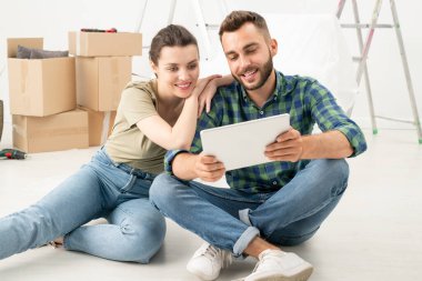 Gülümseyen genç çift yeni bir dairede oturmuş tablet kullanarak internetten mobilya sipariş ediyorlar.