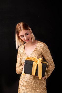 Altın renkli parti elbisesi içinde ne olduğunu tahmin ederken üstünde sarı düğüm olan siyah hediye kutusuna bakan zarif sarışın kadın.