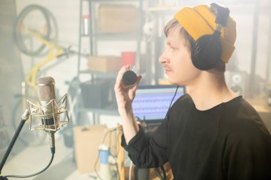 Kulaklıklı genç bir müzisyen mikrofonun önünde oturmuş garajda yeni müzikler kaydediyor.