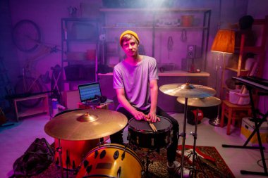 Giyim kuşamında ciddi bir genç ellerini bagetlerle davulun tepesine koyup garajda prova yapıyor.