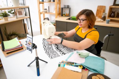 Tekerlekli sandalyede sakat bir kadın evde dikiş makinesinin başında oturmuş çevrimiçi izleyiciler için canlı yayın uzmanlığı yapıyor.