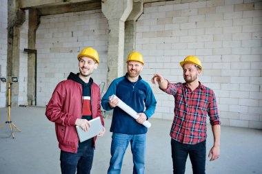 Happy young engineer in hardhat pointing forwards while showing his two colleagues one of walls inside unfinished building at meeting clipart