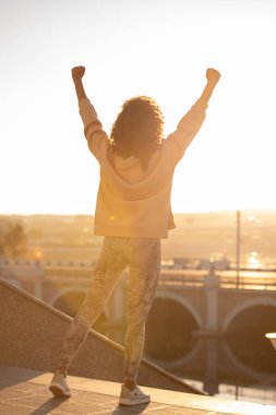 Külotlu çorap ve kapüşonlu genç bayanın kollarını havada tutarken başarı ve memnuniyeti ifade edişinin arka planı.