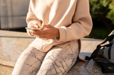 Çağdaş genç kadın milenyumu kapüşonlu ve taytlı açık hava eğitiminden sonra mermer fayansların üzerinde oturuyor ve alet kullanıyor.