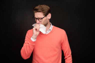 Siyah arka planda durup peçeteye hapşıran gözlüklü hasta genç adam, virüs konsepti.