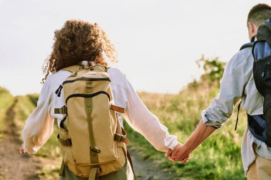 Kıvırcık saçlı, sırt çantası taşıyan, kocasıyla el ele tutuşan ve kırsal yolda ilerleyen genç bir kadın.