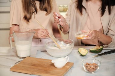 Genç bir kadın sütle cam kaseye bal damlatırken kızı da mutfak masaları ile ahşap kaşıkla dondurma malzemelerini karıştırıyor.