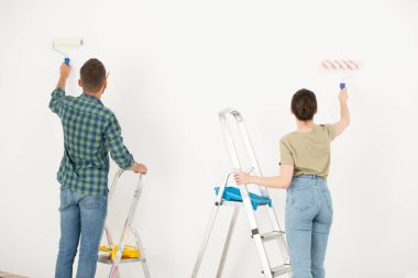 Eski odalarını değiştirirken boya silindiri kullanarak duvarı beyaza boyayan genç çiftin dikiz görüntüsü.