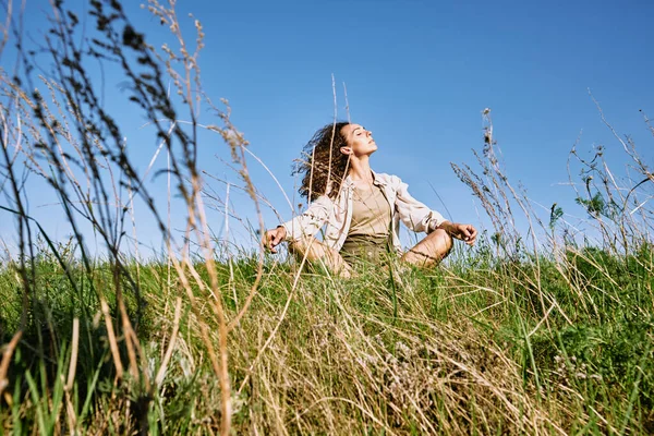 在阳光明媚的日子里 年轻而悠闲的女人坐在碧绿的草地上 长着长长的波浪般的长发 双腿交叉 享受着悠闲的休息 — 图库照片